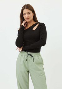 Zwart lang mouwtop met een ribbeltextuur, voorzien van een uitsnijding op de linker schouder, gecombineerd met lichtgroene broek met aantrekkoord.