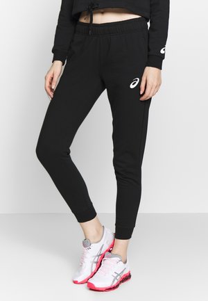 Femme portant un jogging noir ASICS et des chaussures de course blanches avec des semelles roses, debout devant un fond blanc uni.
