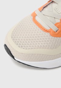 Beżowy sneaker z siateczkową cholewką, pomarańczowymi akcentami i teksturowanymi nakładkami. Biała podeszwa z czarnym wzorem trakcyjnym. Subtelne zakrzywione elementy designu.