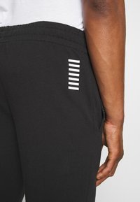 Close-up van zwarte broek met elastische taille en zeven horizontale witte strepen nabij de zak, gedragen door een persoon met een bruine huid.