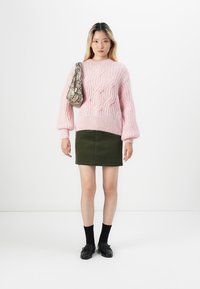 Suéter rosa de malha com padrões de cabos texturizados, combinado com uma saia mini em veludo cotelê verde e loafers pretos. Os acessórios incluem uma mala estampada.