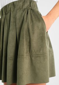 Gonna verde oliva realizzata in morbido materiale simile a suede, con design plissettato, vita elastica e tasche laterali. Presenta una forma leggermente svasata.