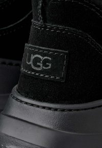 Stivaletto alla caviglia in camoscio nero con suola in gomma, caratterizzato da una toppa con il logo UGG, cuciture a contrasto e punta arrotondata. Dettaglio della superficie testurizzata visibile.