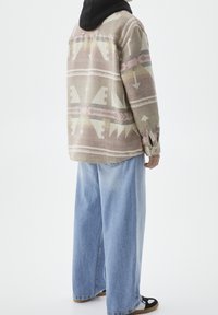 Camicia multicolore con motivo a frecce e forme geometriche, realizzata in un tessuto spesso, indossata su jeans leggeri di colore blu chiaro e sandali neri.