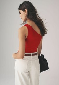 Czerwona ribbed top na ramiączkach z asymetrycznym dekoltem, zestawiona z białymi spodniami o wysokim stanie i brązowym paskiem. Czarna torba trzymana w jednej ręce.