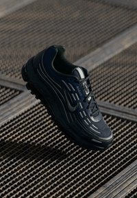 Tamno plavi i crni atletski tenisice s reflektirajućim naglascima, Nike logoom i detaljnim potplatom, smještene na teksturiranoj metalnoj rešetkastoj površini.