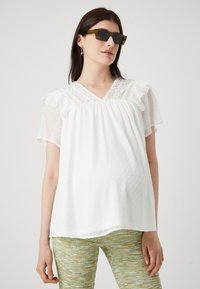 Balta blouse ar punktveida tekstūru, mežģīņu akcentiem un saplacinātām īsajām piedurknēm. Pāris ar rakstainām zaļām biksēm. Uzliktas saulesbrilles.
