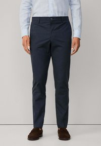 Man i slimfit marinblå byxor, ljusblå randig skjorta med knappar och mörkbruna mockaloafers, stående mot en enfärgad beige bakgrund.