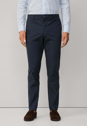 Mann in schmal geschnittenen navyblauen Hosen, hellblau gestreiftem Button-Down-Hemd und dunkelbraunen Wildleder-Slippern, steht vor einem einfarbigen beigen Hintergrund.
