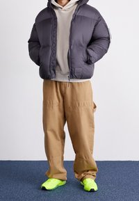 Puffjacke in Dunkelgrau mit einem Vorderreißverschluss, kombiniert mit beigen Zugbandhosen und leuchtend gelben Sneakern. Material aus Baumwollmix.