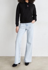 Černá mikina s kapucí a přední kapsou, malým logem. Světle modré rozšířené džíny s čistým designem. Modelka má na sobě černé podpatky.