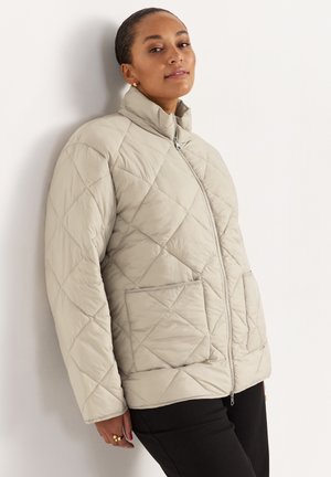 Frau lehnt an der Wand, trägt eine beige gesteppte Jacke mit Fronttaschen und schwarze Hosen.