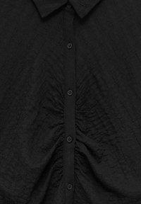 Chemise noire à boutons avec un tissu texturé. Elle présente un col, des fronces verticales sur le devant et cinq boutons ronds.