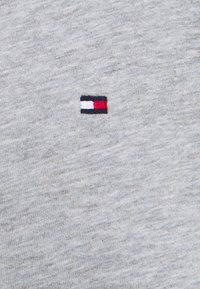 Grauer Baumwollstoff mit einer subtilen Textur. Verfügt über ein kleines gesticktes Logo mit roten, weißen und blauen Elementen auf der linken Seite.