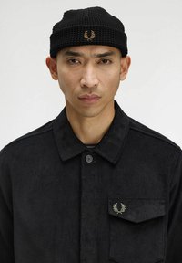 Uomo che indossa una giacca nera a coste con colletto abbottonato e berretto di maglia nero con loghi di un alloro ricamati in oro.