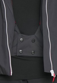 Giacca nera con zip frontale, caratterizzata da una texture liscia, foderatura interna rossa e un accento con chiusura a scatto al centro.