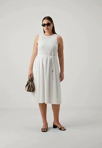 Žena u bezrukavoj bijeloj midi haljini s remenom, crnim sunčanim naočalama, crnim sandalama na visoku petu s zlatnim lancem i drži metalik bež torbicu.