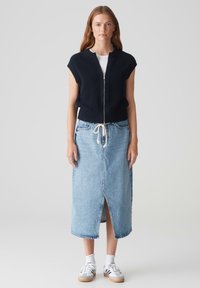 Canotta a coste blu navy con maniche corte, chiusura con zip, abbinata a una gonna in denim chiaro a vita alta con uno spacco frontale e sneakers bianche casual.