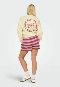 Sweatshirt crème avec le texte rouge "NEW YORK CITY BROOKLYN 1993" dans le dos. Associé à un short rayé bordeaux et blanc, porté avec des baskets blanches.