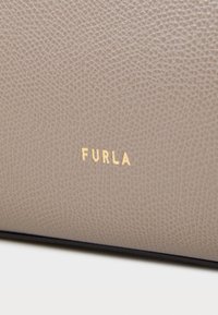 Jasno beżowa skórzana powierzchnia o teksturowanym wykończeniu. Złote, wytłoczone logo "FURLA" umieszczone na środku materiału. Gładkie krawędzie, minimalistyczny design.