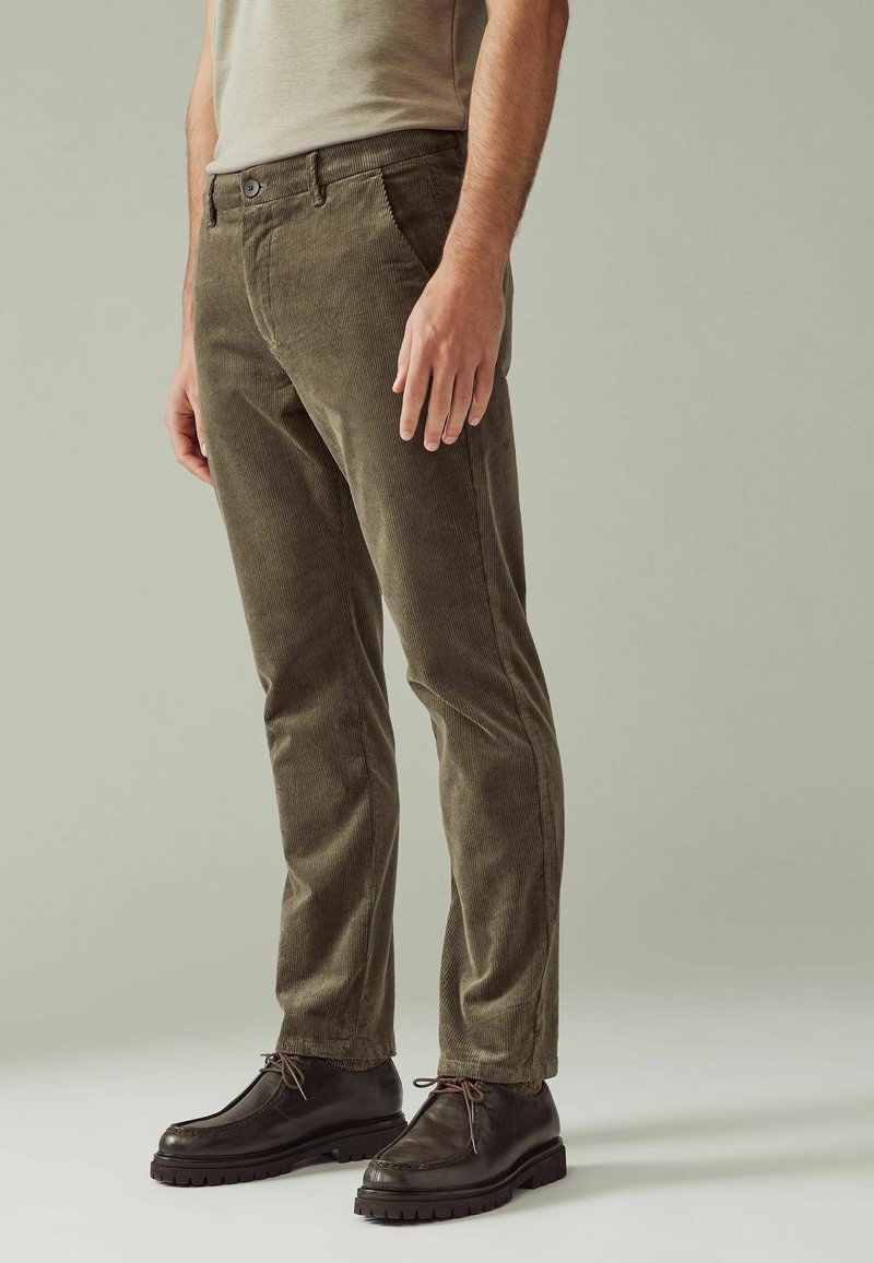Homme portant un pantalon en velours côtelé vert olive et des chaussures à lacets marron foncé, debout devant un fond uni gris clair.