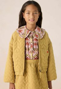 Veste matelassée jaune moutarde avec des motifs en forme de cœur, associée à un chemisier à fleurs roses avec un col ruffled et une jupe assortie.
