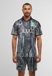 Mann in schwarzem und grauem gemustertem Sporttrikot und Shorts mit Logos, der vor einem einfarbigen hellgrauen Hintergrund steht.