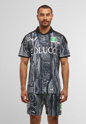 Uomo che indossa una maglia sportiva a motivo nero e grigio e pantaloni corti con loghi, in piedi contro uno sfondo grigio chiaro uniforme.