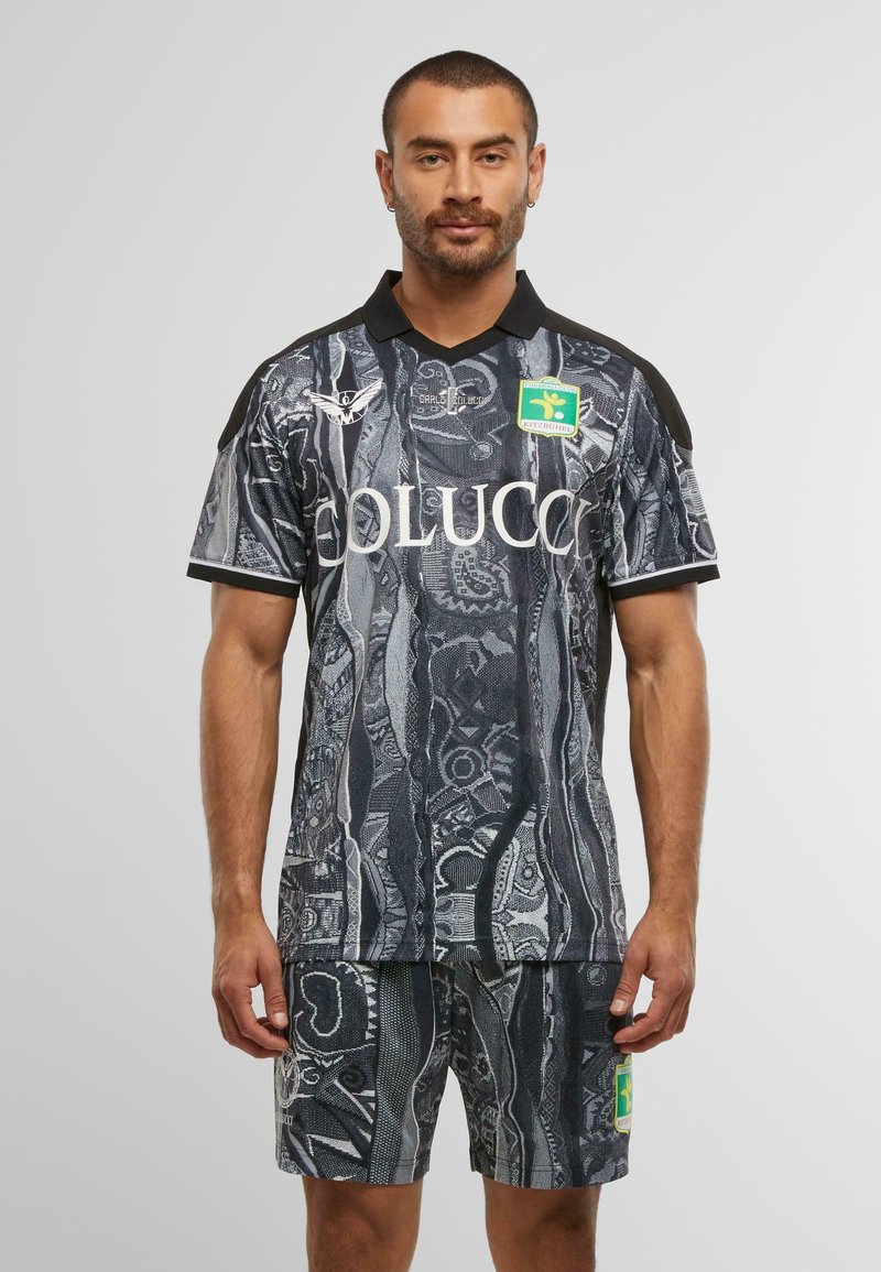 Mann in schwarzem und grauem gemustertem Sporttrikot und Shorts mit Logos, der vor einem einfarbigen hellgrauen Hintergrund steht.