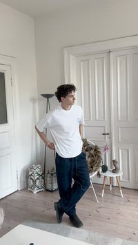Camiseta branca com um logo azul, calças largas escuras e botas pretas. Quarto claro e neutro com chão em madeira e móveis suaves.