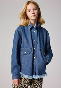 Camicia in denim di blu medio con chiusura a bottoni, due tasche sul petto e orlo sfrangiato. Abbinata a pantaloni a stampa leopardata.