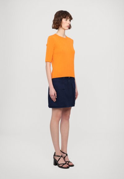 Oranžna rebrasta kratka majica s kratkimi rokavi v kombinaciji z mornarsko modrim denim mini krilom, ki ima sprednje žepke in ravno silhueto. Črne sandale z visokimi petami in trakci.