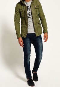 Oliwkowa kurtka z kapturem z wieloma kieszeniami, noszona na szarym t-shircie z grafiką, w połączeniu z ciemnymi wąskimi dżinsami i czarnymi trampkami.
