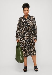 Robe à motifs noirs et beiges avec un col, des manches longues et une ceinture à la taille. Associée à un sac à main vert et des sandales à plateforme noires.