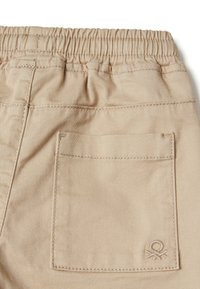 Beżowe bawełniane spodnie z elastycznym pasem, tylną kieszenią oraz małym haftowanym logo na kieszeni. Gładka tekstura.