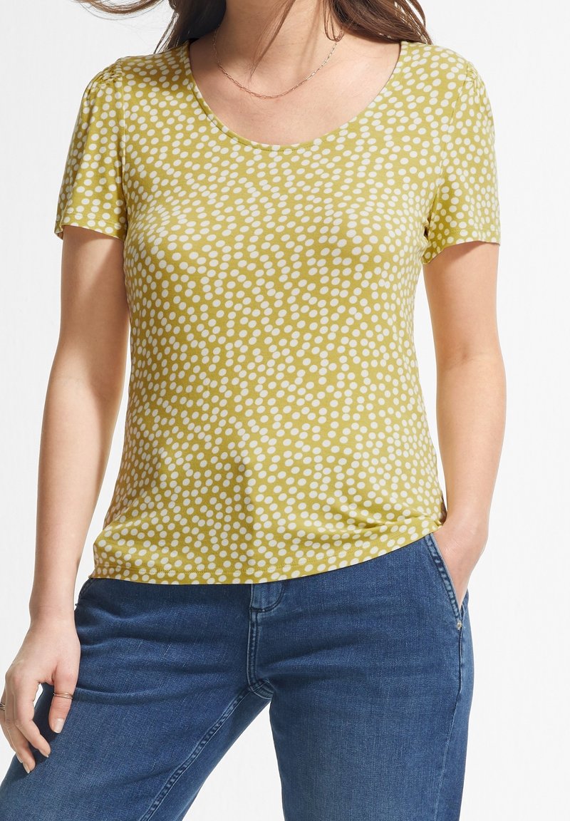 Femme portant un haut jaune à manches courtes avec des motifs à points blancs et un jean bleu, debout avec une main dans la poche, sur fond blanc.