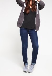 Sudadera con capucha de cremallera en gris oscuro con cordones blancos sobre una camiseta negra, combinada con jeans azules y zapatillas grises. Modelo de pie con los brazos levantados.
