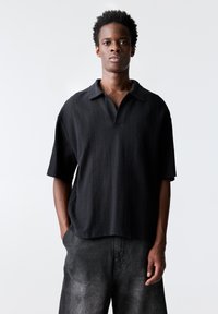 Polo shirt nera a maniche corte con un design a maglia testurizzata e colletto classico. Indossato con pantaloni corti scuri e sbiaditi.