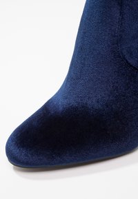 Gros plan sur la pointe d'une chaussure à talon haut en velours bleu marine avec un design pointu sur fond blanc.