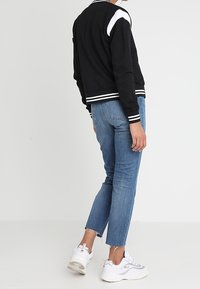 Black bomber jacket with white accents, ribbed cuffs and hem. Paired with light blue, cropped jeans and white sneaker shoes. Back view.
