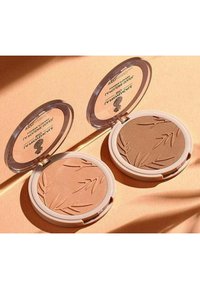 Deux boîtiers ronds ouverts de poudre bronzante ; un beige clair avec un motif bambou, un brun foncé, tous deux avec des couvercles transparents et des bords blancs.