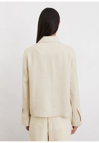 Licht beige, op maat gemaakte jas van een linnenmix. Heeft een ongelijke kraag, lange mouwen met openingen en een minimalistisch ontwerp zonder versieringen.