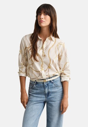 Femme portant une chemise blanche à boutons avec des motifs de chaînes dorées, un jean bleu clair et une ceinture blanche avec une boucle dorée, debout devant un fond blanc.