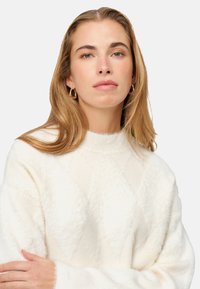 Pull en maille duveteuse de couleur crème avec un motif en losanges texturé, un col montant et des manches larges, porté par une personne aux cheveux longs.