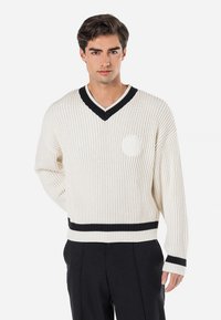 Cream ribbed sweater with black V-neck and trim. Features a round logo on the chest and black accents on the cuffs and hem.