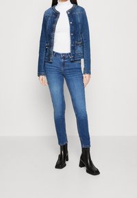 Mujer vestida con una chaqueta de mezclilla azul, un cuello de tortuga blanco, jeans azules y botas de tobillo negras con tacones, de pie contra un fondo liso.