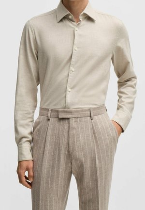 Mann trägt beige langärmliges Hemd mit Knöpfen, in hoch taillierte hellbraune Nadelstreifenhose mit Gürtelschlaufen gesteckt.
