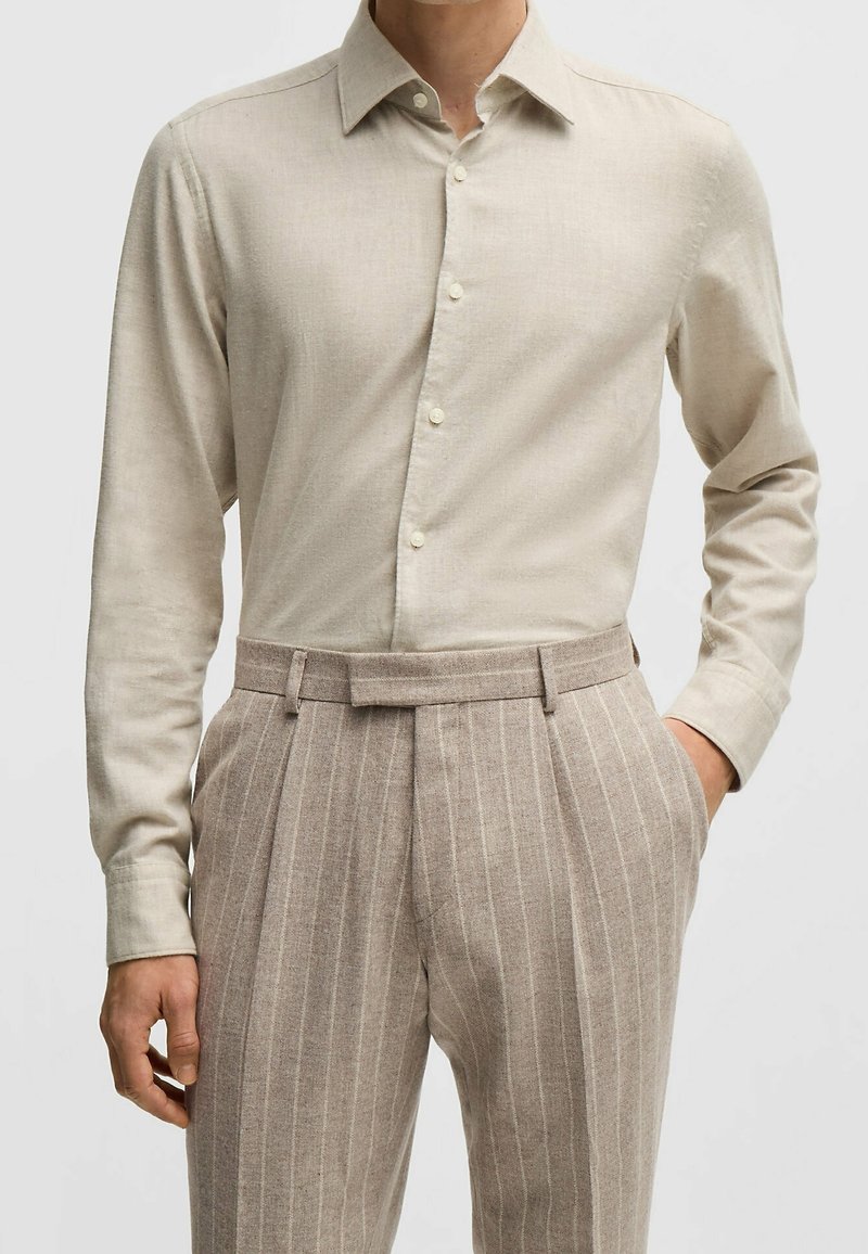 Homme portant une chemise beige à manches longues boutonnée, rentrée dans un pantalon à fines rayures marron clair taille haute avec passants de ceinture.
