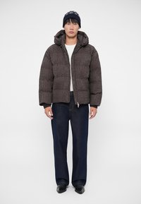 Brūns pūkains jaka ar quilted tekstūru un kapuci, apvienota ar tumšiem džinsu biksēm. Galvā adīts beanie ar rakstainu jostu.