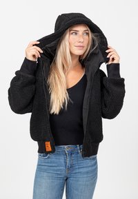 Sweat à zip noir en matériau duveteux avec une capuche doublée, des poignets côtelés et une étiquette orange sur l'ourlet. Porté par-dessus un haut noir.